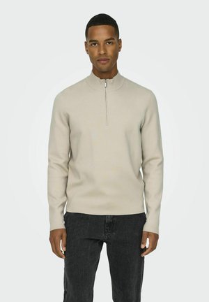 Light beige, zip-neck sweater with long sleeves, ribbed collar, and cuffs, paired with dark gray jeans. Smooth texture and fitted silhouette.