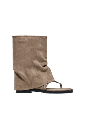 Hoge beige suède sandaal-laars hybride met een zwarte platte zool en een teenbandje op een witte achtergrond.