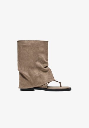 Sandale-bottine hybride en daim beige clair avec une semelle plate noire et une bride en lanière sur un fond blanc.