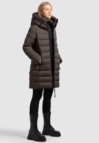 Brązowa puchowa kurtka z wysokim kołnierzem, poziomym pikowaniem, zamkiem błyskawicznym z przodu i bocznymi kieszeniami, w połączeniu z czarnymi butami na sznurówki.