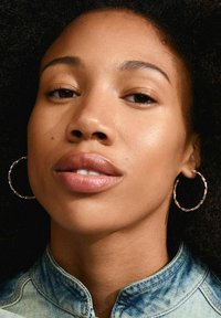 Primer plano de la cara de una persona con cabello oscuro y rizado. Lleva pendientes grandes de aro de plata retorcidos y una chaqueta de mezclilla con cuello.