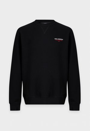 Un mannequin masculin se tient devant un fond blanc, vêtu d’un sweat-shirt noir avec logo, d’un pantalon noir et de baskets noires en toile.