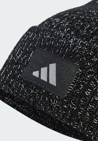 Svart stickad mössa med en strukturerad mönster och vita fläckar. Inkluderar en rektangulär svart etikett med en silverfärgad grafisk logotyp.