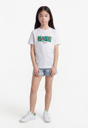 Κορίτσι που στέκεται φορώντας ένα λευκό μπλουζάκι Nike με πολύχρωμο λογότυπο, ανοιχτόχρωμα τζιν σορτς και λευκά αθλητικά παπούτσια με πράσινες λεπτομέρειες.