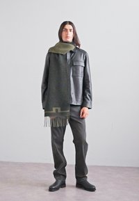 Veste en cuir noir avec une poche poitrine, écharpe en laine verte avec des franges, pantalon gris et chaussures noires, le tout sur un fond neutre.
