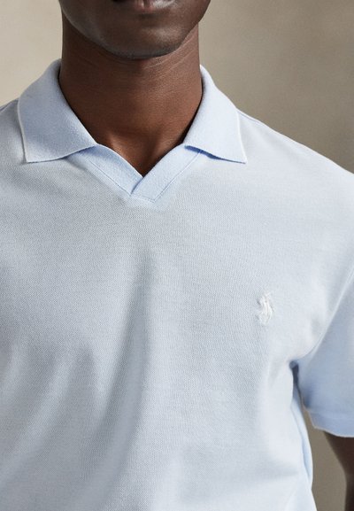 Lyseblå poloshirt laget av teksturert stoff, med V-hals og brodert logo på venstre bryst.