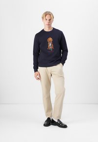 Felpa blu navy con un grafico di un cane sul davanti, abbinata a pantaloni beige e scarpe nere. Design semplice, texture morbida, vestibilità casual.