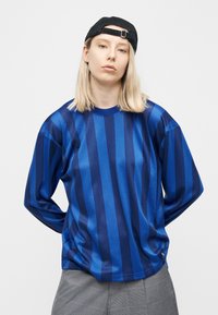 Νεαρός με ξανθά μαλλιά που φοράει μπλε ριγέ μακρυμάνικο πουκάμισο, γκρι παντελόνι και μαύρο καπέλο με το γείσο προς τα πίσω, στέκεται με τα χέρια πίσω από την πλάτη.