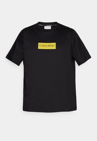 Černé bavlněné triko s žlutým obdélníkovým logem s textem "Calvin Klein" a "ZALOŽENO 1968". Klasický design s kulatým výstřihem.