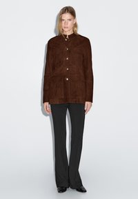 Veste en daim marron avec un col montant, des boutons dorés et deux poches avant ; associée à un pantalon noir évasé et des chaussures noires pointues.