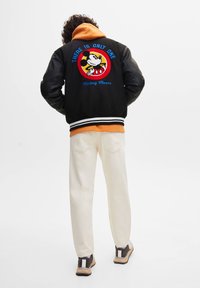 Veste universitaire noire avec manches en cuir, ornée d'un motif brodé de Mickey Mouse en rouge, jaune et bleu au dos. Ourlet à rayures blanches.