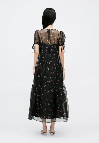 Rochie maxi neagră cu flori, din țesătură transparentă, mâneci bufante și fustă cu volane. Are închidere cu fundă la spate și modele delicate de flori.