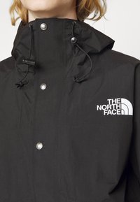 Personne portant une veste noire North Face avec un col montant, des cordons ajustables et des boutons-pression en métal visibles à l'avant.