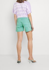 Shorts en denim vert menthe avec une coupe ajustée, cinq poches et des coutures décoratives à l'arrière. Associés à un t-shirt violet tie-dye.