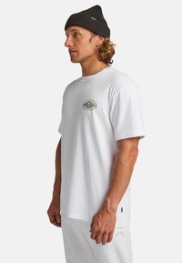 Camiseta blanca de algodón con mangas cortas, escote redondo y un gráfico circular multicolor en el pecho. Llevada con pantalones de chándal grises.