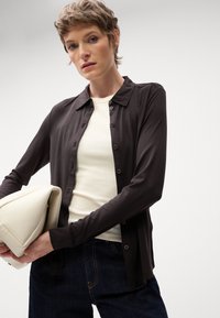 Mørkebrun button-up skjorte med krave, bæret over en lys beige ribbet top. Modellen holder en cremefarvet taske med en glat overflade.