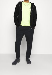 Felpa nera con zip sopra una t-shirt verde neon, abbinata a pantaloni jogger scuri e sneakers nere con texture. Dettaglio accentato in catena argentata.