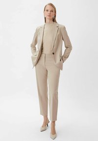 Beżowy, dopasowany garnitur z jednorzędową marynarką, dopasowanymi spodniami oraz jasną, prążkowaną bluzką; elegancki design z szpiczastymi, beżowymi szpilkami.