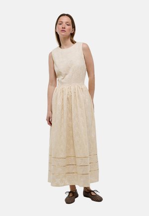 Femme debout portant une robe midi fleurie crème sans manches avec une taille froncée et des chaussures à lacets sombres à motifs.