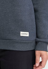 Mørk grå hettegenser med en myk tekstur. Har ribbestrikkede mansjetter og kant, med et rektangulært hvitt merke som viser merket "oxmo."