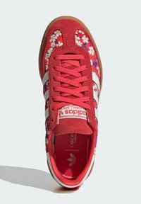 Zapatilla Adidas roja con rayas blancas, paneles de tela con patrón floral, detalles de ante y suela de goma, vista desde arriba sobre un fondo claro.