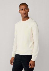 Mann mit kurzen Haaren, der einen schlichten weißen Rundhalsausschnitt-Pullover und dunkle Hosen trägt, und leicht zur Seite blickt, vor einem neutralen Hintergrund.