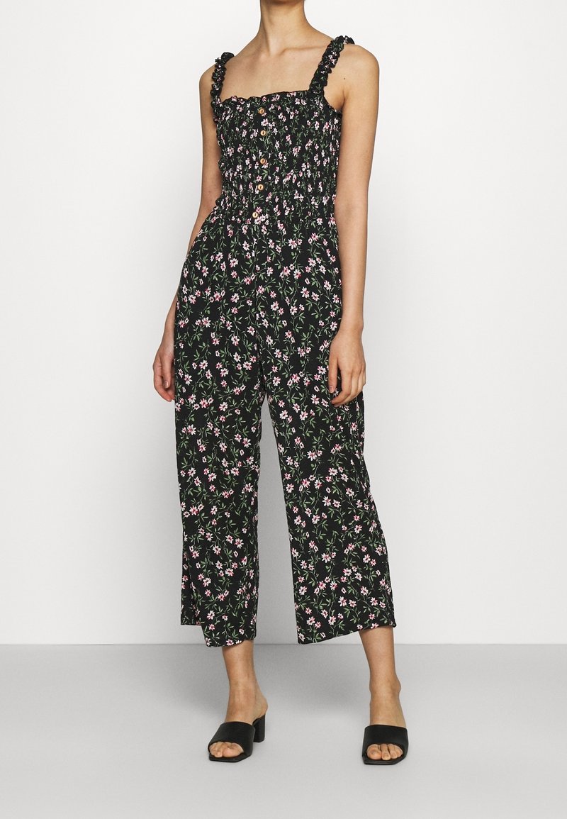 Femme portant une combinaison noire sans manches avec imprimé floral rose et blanc et bretelles à volants, associée à des sandales noires à talons ouvertes.