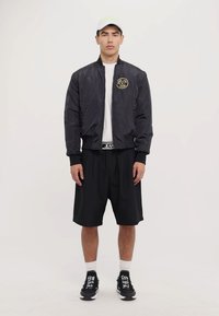 Svart bomberjacka med guldemblem, vit t-shirt, svarta shorts, svarta sneakers och en ljus färgad keps. En enkel, modern atletisk outfit.