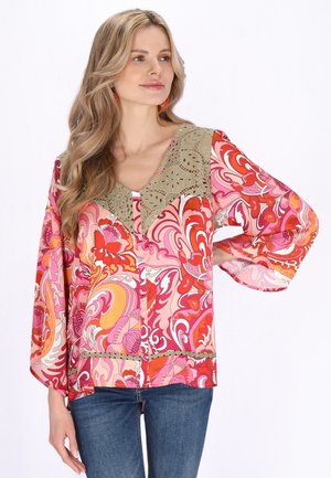 Kvinde iført en flerfarvet, blomsterprintet bluse med vide ærmer og blonde detaljer, kombineret med blå jeans, ser til siden.