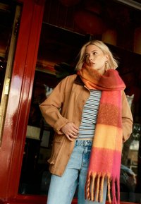 Braune knöpfbare Jacke mit einem dunklen Kragen, gestreiftem Hemd darunter und einem lebhaft orange, rosa und gelb karierten Schal mit Fransen.