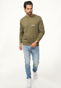Olive green sweatshirt with a round neckline and ribbed cuffs, paired with light blue jeans and white sneakers. Features a small logo on the chest.