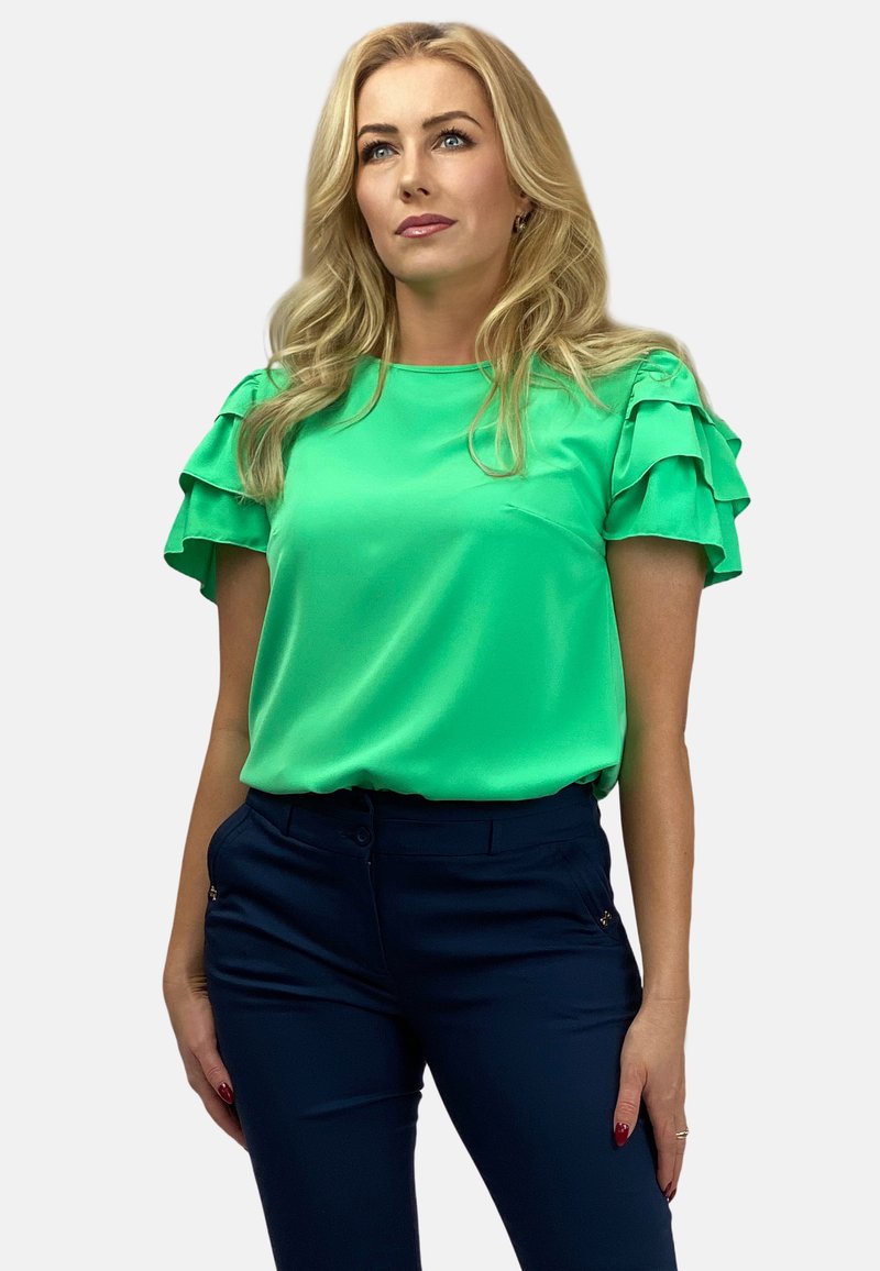 Zielona satynowa bluzka z falbaniastymi rękawami, w zestawieniu z granatowymi spodniami. Materiał ma gładką teksturę i luźny krój.