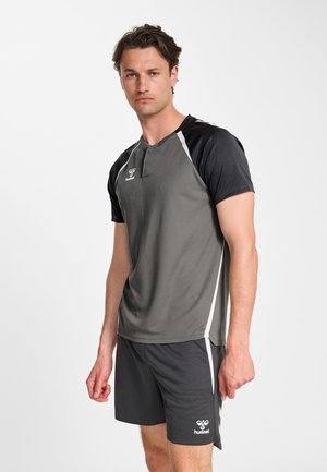 Uomo con abbigliamento sportivo grigio e nero, in piedi con un'espressione neutra su uno sfondo bianco uniforme.