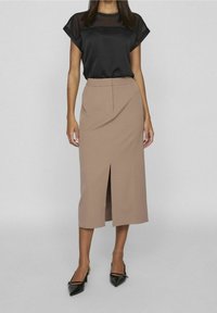 Blouse noire transparente à manches courtes, associée à une jupe crayon beige longueur genou avec une fente à l'avant, portée avec des chaussures noires à bouts pointus.