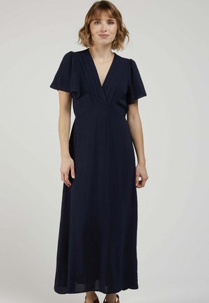 Femme aux cheveux courts portant une robe maxi bleu marine à manches courtes et col en V, sur un fond gris clair uni.