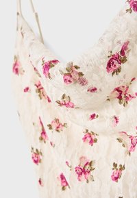 Robe en tissu en dentelle florale avec des roses brodées roses et vertes. Texture douce, design à col drapé et fines bretelles. Fond crème.