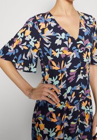 Kobieta w granatowej sukience w wielokolorowy kwiatowy wzór, z dekoltem w serek i krótkimi falbankami, ręka na biodrze, z srebrnymi pierścionkami i bransoletką.