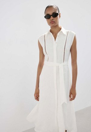 Žena nosi bijelu haljinu-košulju bez rukava s remenom, okomito rezanim čipkastim detaljima i crnim sunčanim naočalama mačje oči na jednostavnoj pozadini.