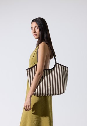 Femme aux longs cheveux foncés portant une robe sans manches couleur moutarde, portant un grand sac cabas rayé marron et beige sur l'épaule.