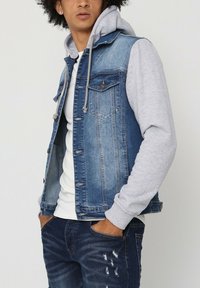 Veste en denim avec des manches de hoodie gris, délavage bleu clair à moyen, deux poches poitrine, boutons en métal, détails usés, portée avec un jean noir.