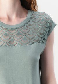 Krátký rukáv top v měkké zelené látce se průhledným, vzorovaným krajkovým potahem, který má vlnité designy podél výstřihu a ramen.