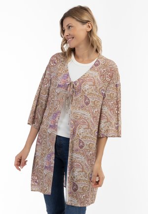 Lang cardigan med paisley-mønster og bændel foran, der har en beige bundfarve, lilla og gyldne detaljer samt brede ærmer, båret over en hvid top.