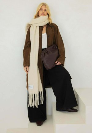 Manteau en suède marron sur un haut blanc, écharpe crème, pantalon noir à larges jambes et sac violet foncé. Frange visible sur l'écharpe. Chaussures marron.