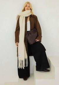Bruine suède lange jas, crème wollen sjaal met franjes, zwarte wijde broekspijpen en een donkerpaarse tas met geplooid ontwerp.