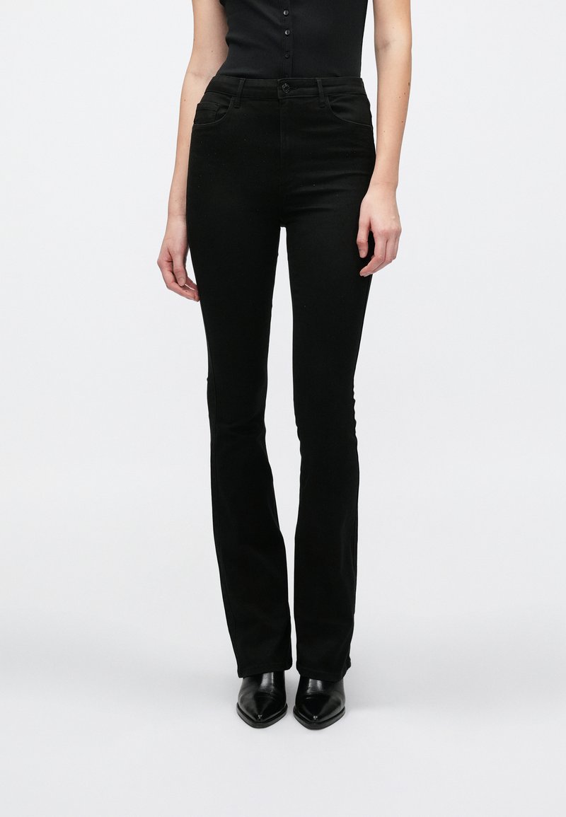 Μαύρο τζιν με φαρδιά καμπάνα και λεία, ελαστική υφή, μέση μεσαίου ύψους, μπροστινές τσέπες και απλή κλείσιμο με κουμπί. Φορεμένο με μυτερά μαύρα παπούτσια.