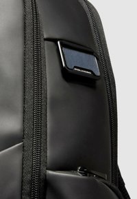 Zaino nero con tessuto testurizzato, superficie liscia e una cerniera prominente. Un'etichetta metallica con il nome del marchio è attaccata.