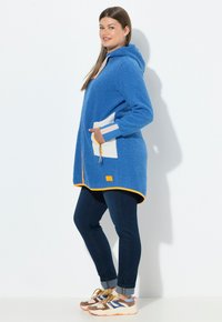 Blaue Fleece-Hoodie mit einer vorderen Kängurutasche, weißen Akzenten, orangefarbenen Details und einstellbarem Kordelzug. Kombiniert mit dunklen Jeans und Sneakers.