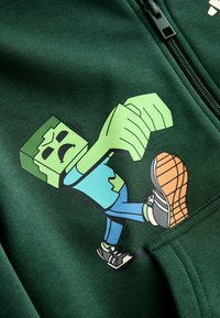 Grüner Sweatshirtstoff zeigt eine Grafik eines Cartoon-Charakters mit einem blockförmigen Kopf, grüner Haut, blauem Hemd und Sneakers, der gerade einen Schritt macht.