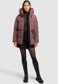 Chaqueta de plumas en un suave malva, con cuello alto, cierre con cremallera en la parte delantera y dos bolsillos laterales. Lleva puesta una blusa y una falda negras. Botas negras hasta la rodilla.