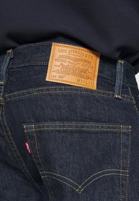 Vaqueros de mezclilla azul oscuro con una etiqueta de cuero marrón en la cinturilla, con costuras amarillas y un diseño clásico de cinco bolsillos.